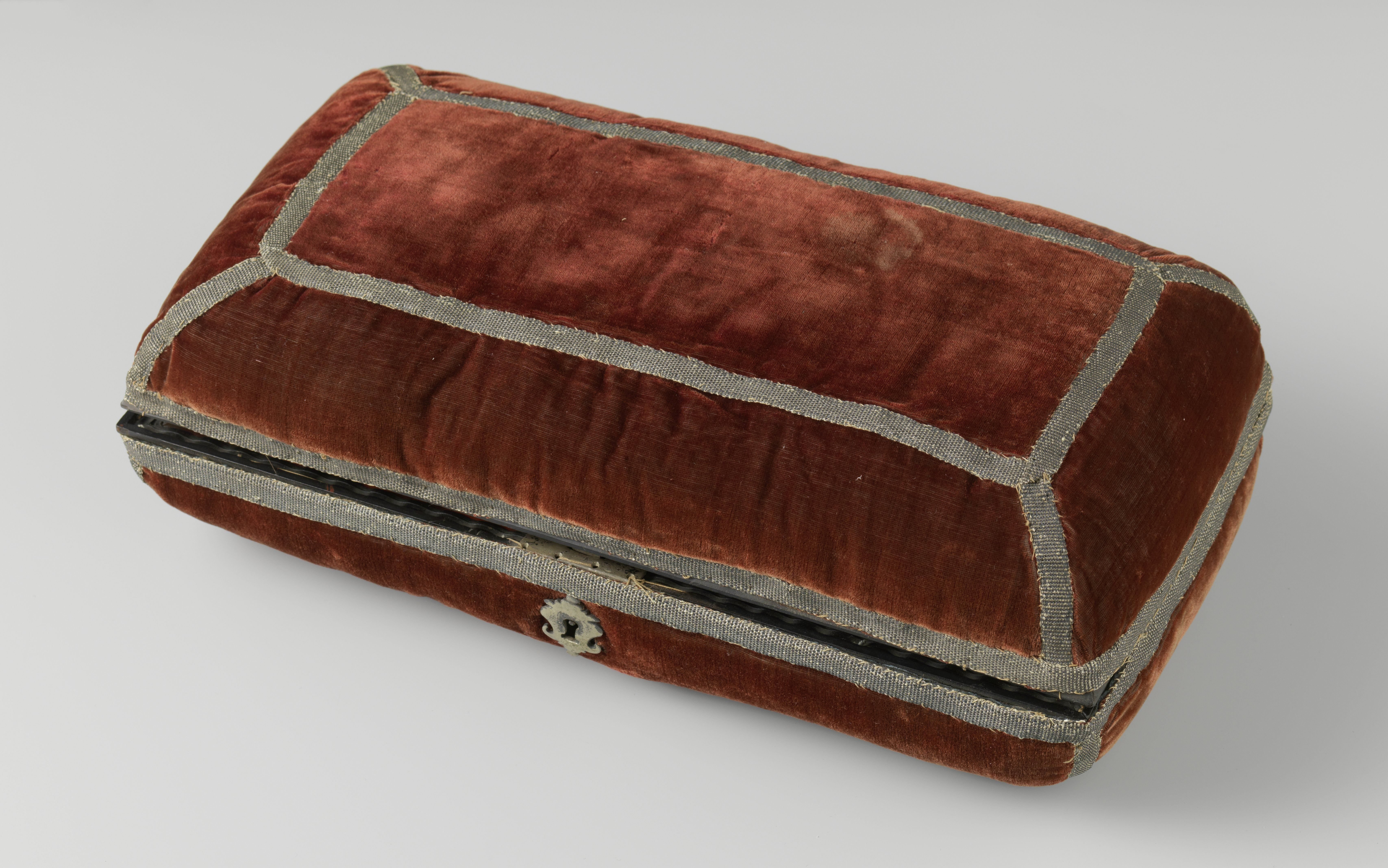 Langwerpig kussen bestaande uit twee gelijke helften, bekleed met rood fluweel versierd met zilveren passement. Binnenwerk van hout belijmd met schildpad en voorzien van compartimenten met beschilderde deksels met rollijsten van ebbenhout. Messing slot middenvoor.