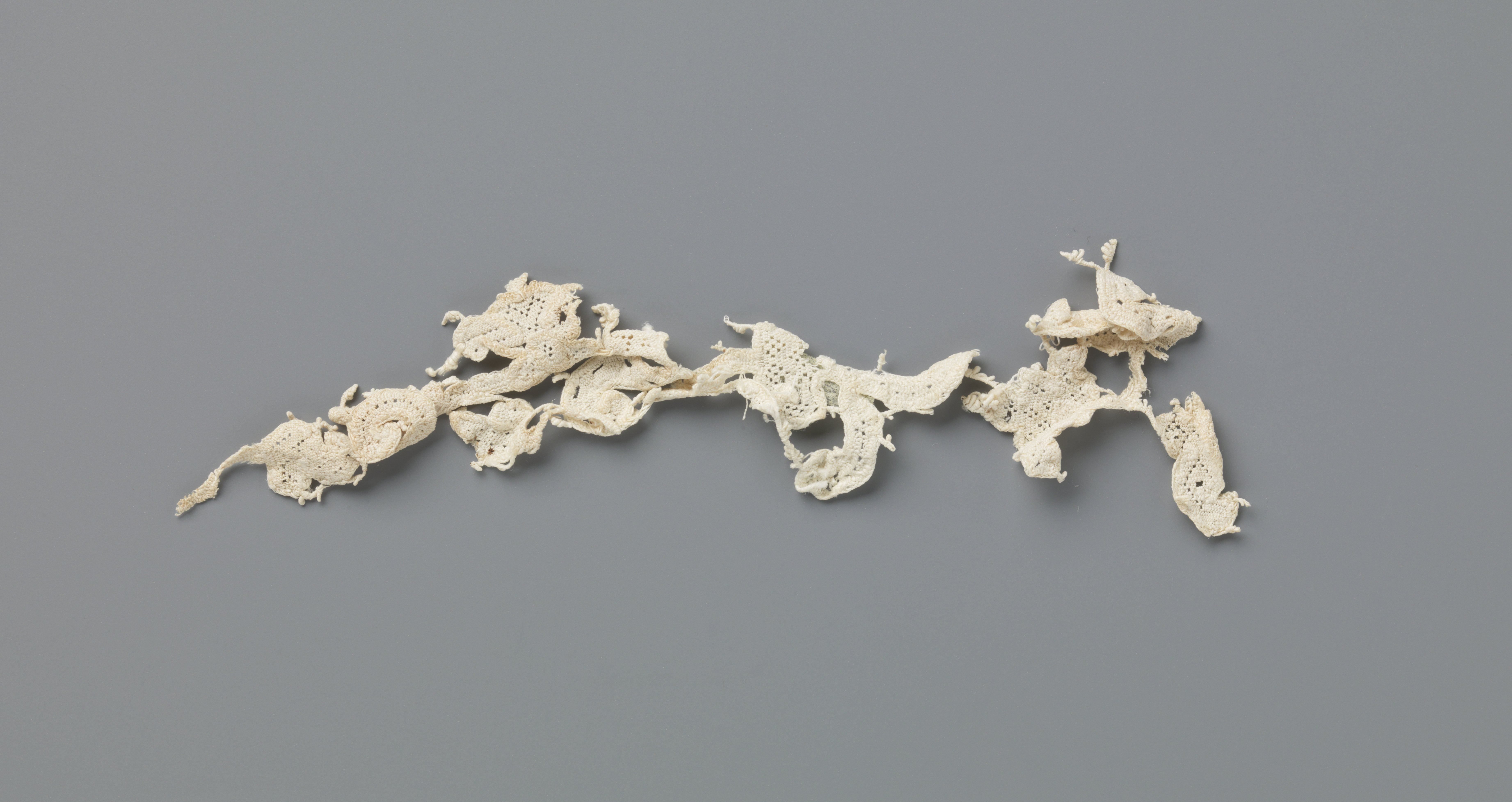 Fragment van een strook naaldkant met krullende ranken en veelsoortige bloemen
