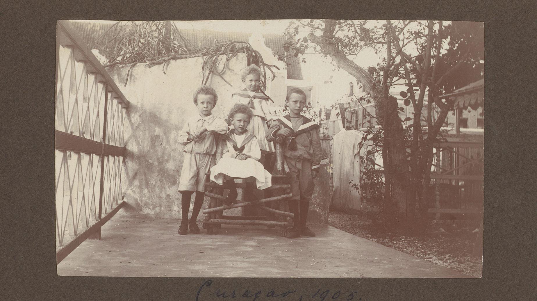 Portret van vier kinderen buiten op een houten bank in Curaçao, vermoedelijk de kinderen van Jan Olphert de Jong van Beek en Donk