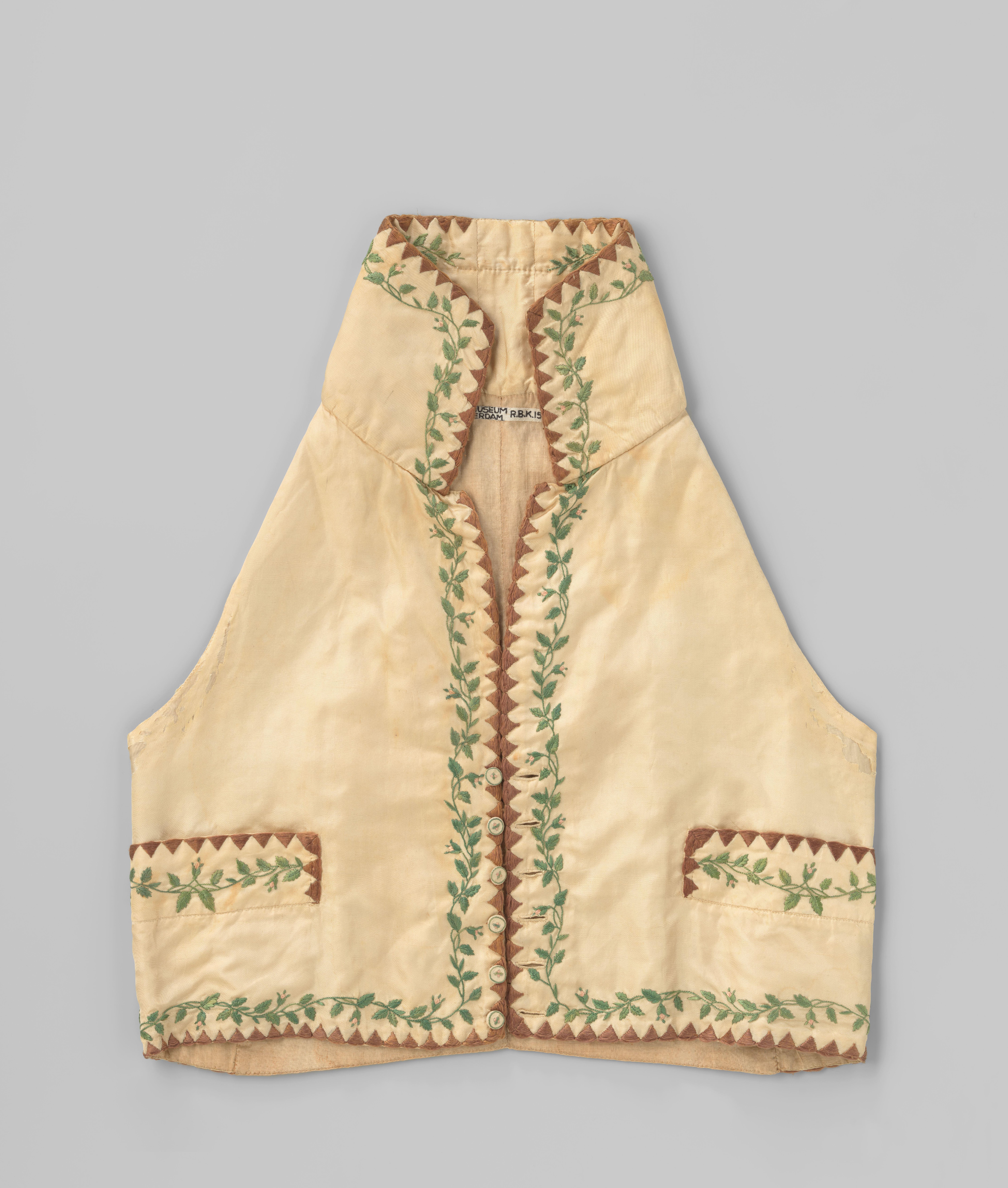 Zeer kort vest met liggende kraag, revers en zes stofknopen van crème zijde, met bruine geborduurde rand van driehoeken en geborduurde groene bladranken met roze knoppen