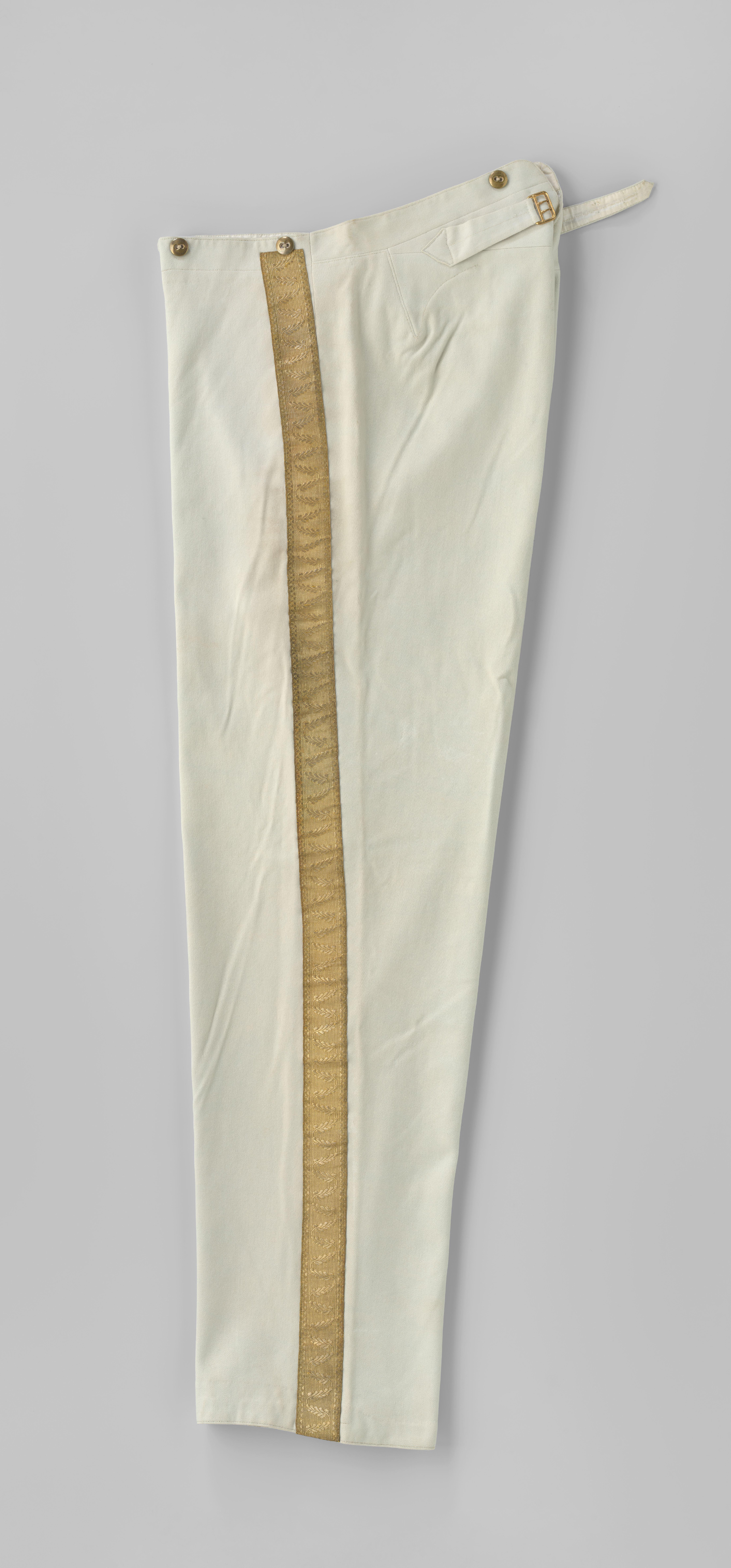 Lange broek van wit laken met koperen knopen, taille met V-vormige split en bandjes met gouden gesp; langs de pijpen afgezet met goudgalon met bladslinger.
