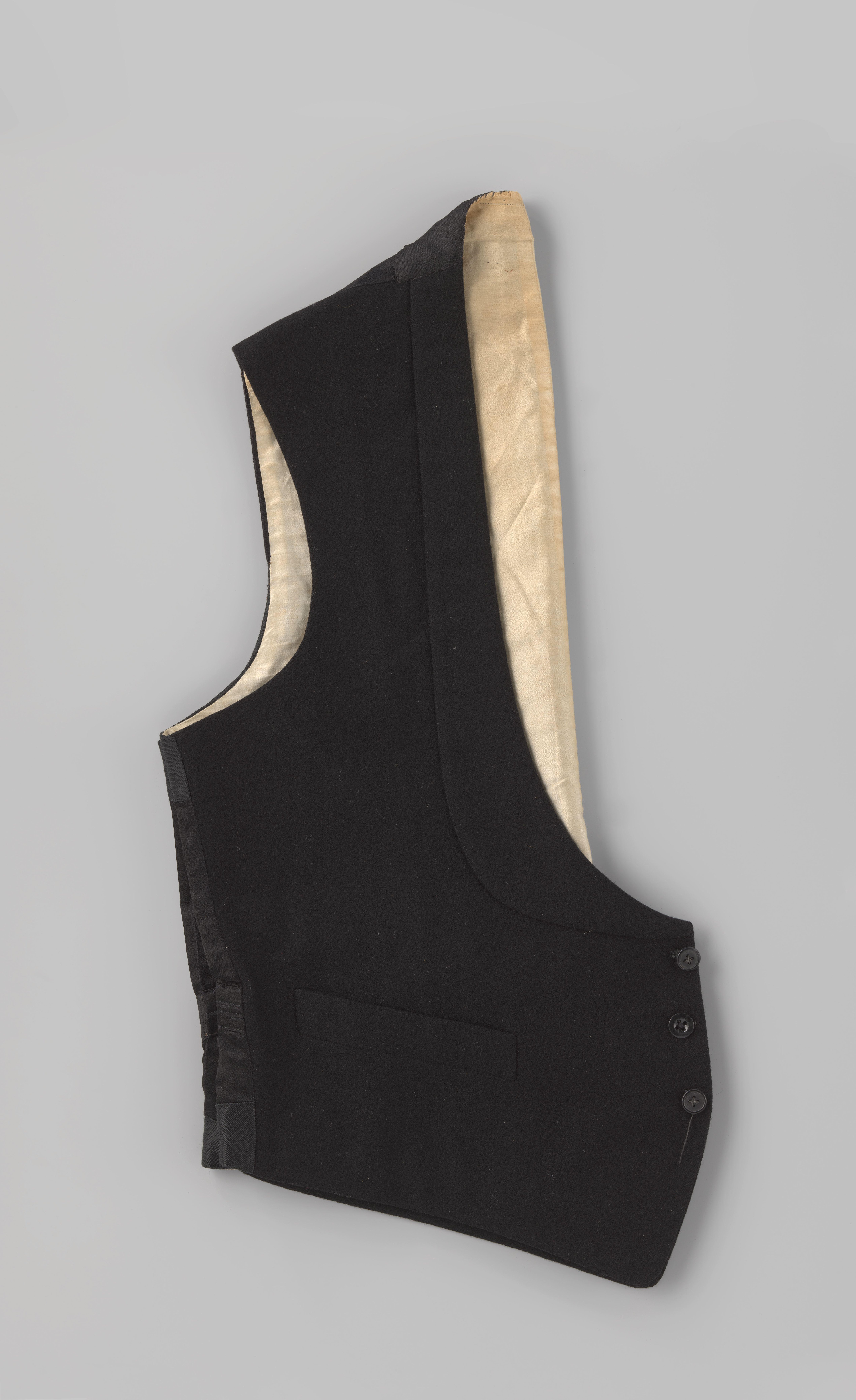 Smokingvest van zwart laken, met diepe sjaalkraag, drie knopen en drie steekzakken; rug van zwart satijn (?) met bandje en gesp en voering van wit katoen