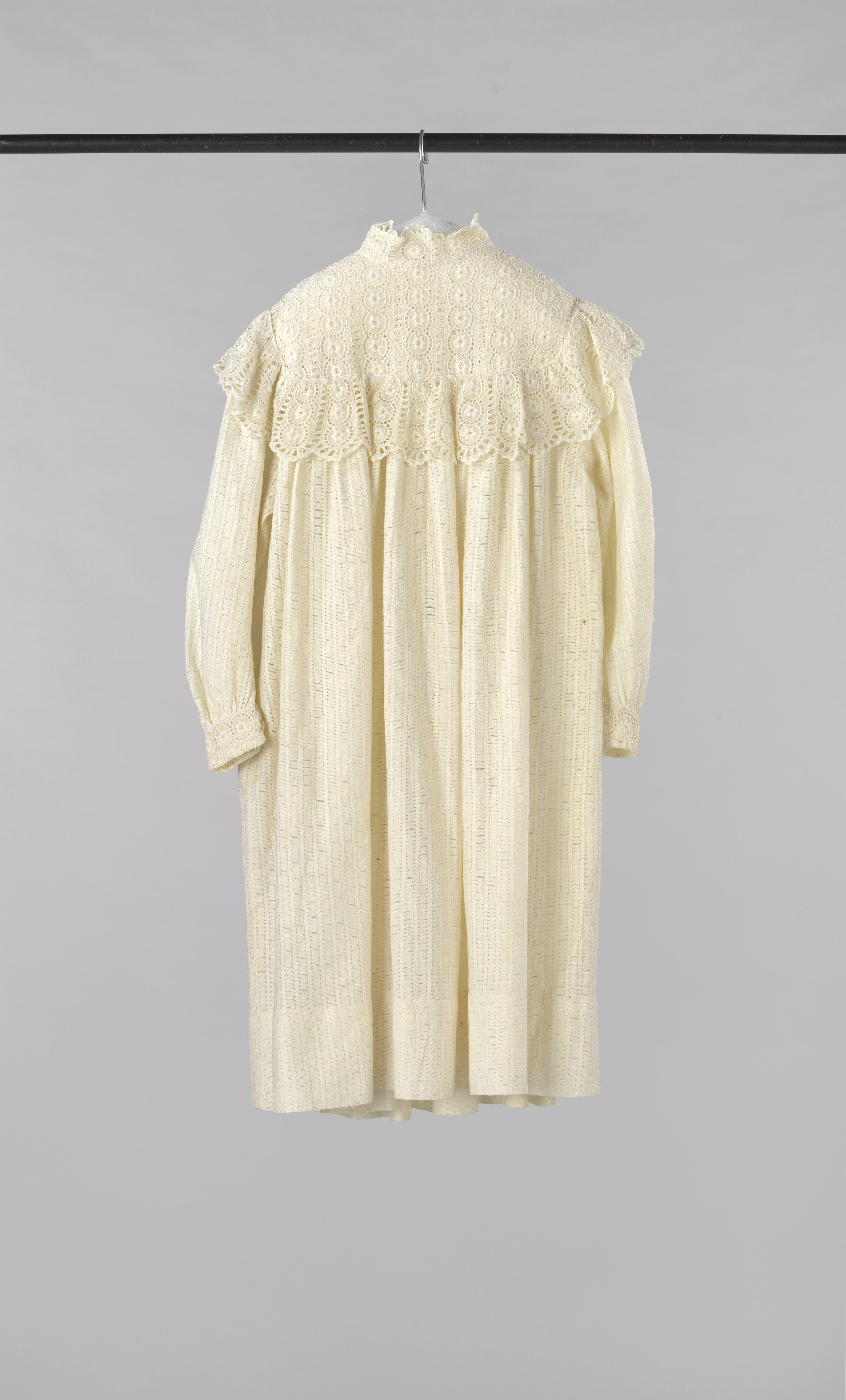 Meisjesjurk met lange mouw, van gedessineerd neteldoek (?) met streepjes- en blokmotiefjes; versierd met pas en strook van randstof met machinal Engels borduurwerk