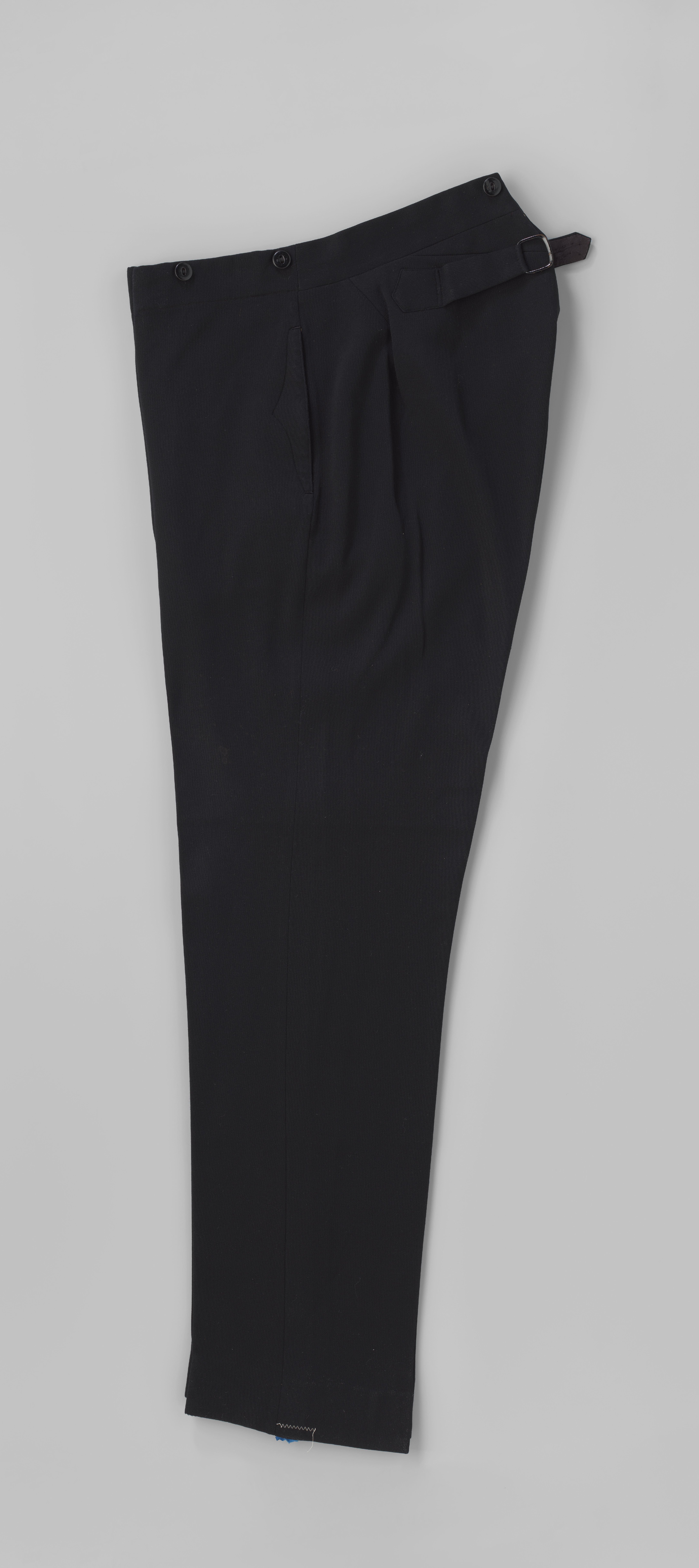 Lange broek van zwart laken, met nauw toelopende pijpen zonder omslag, steekzakken opzij met knoopsluiting, gulp met knopen en zes knopen op de tailleband