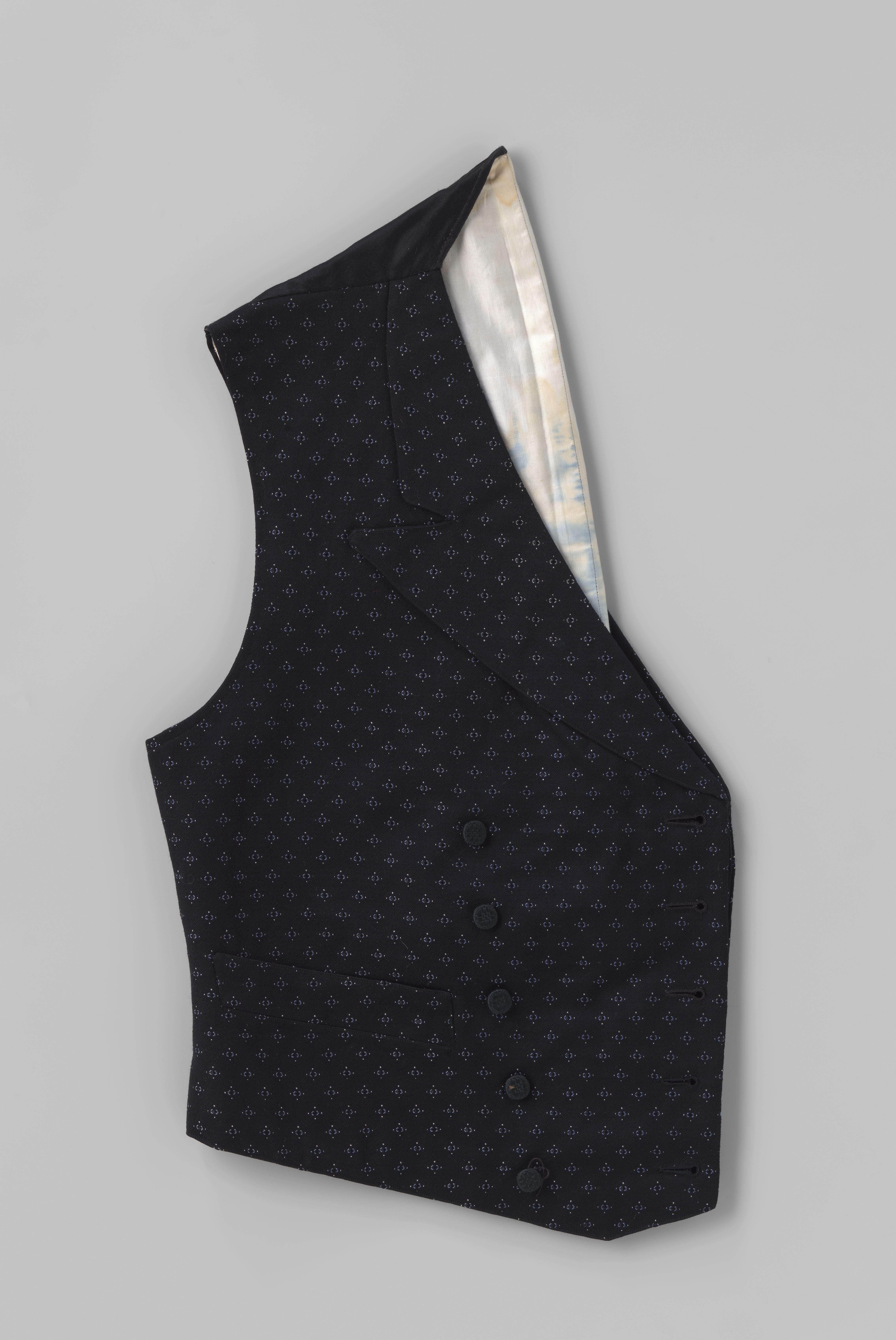 Wintervest van wol, zwart met blauwe figuurtjes met witte stipjes, kort model, de voorpanden van onder in een boog gesneden en met overslag met dubbele rij zwarte passementknoopjes