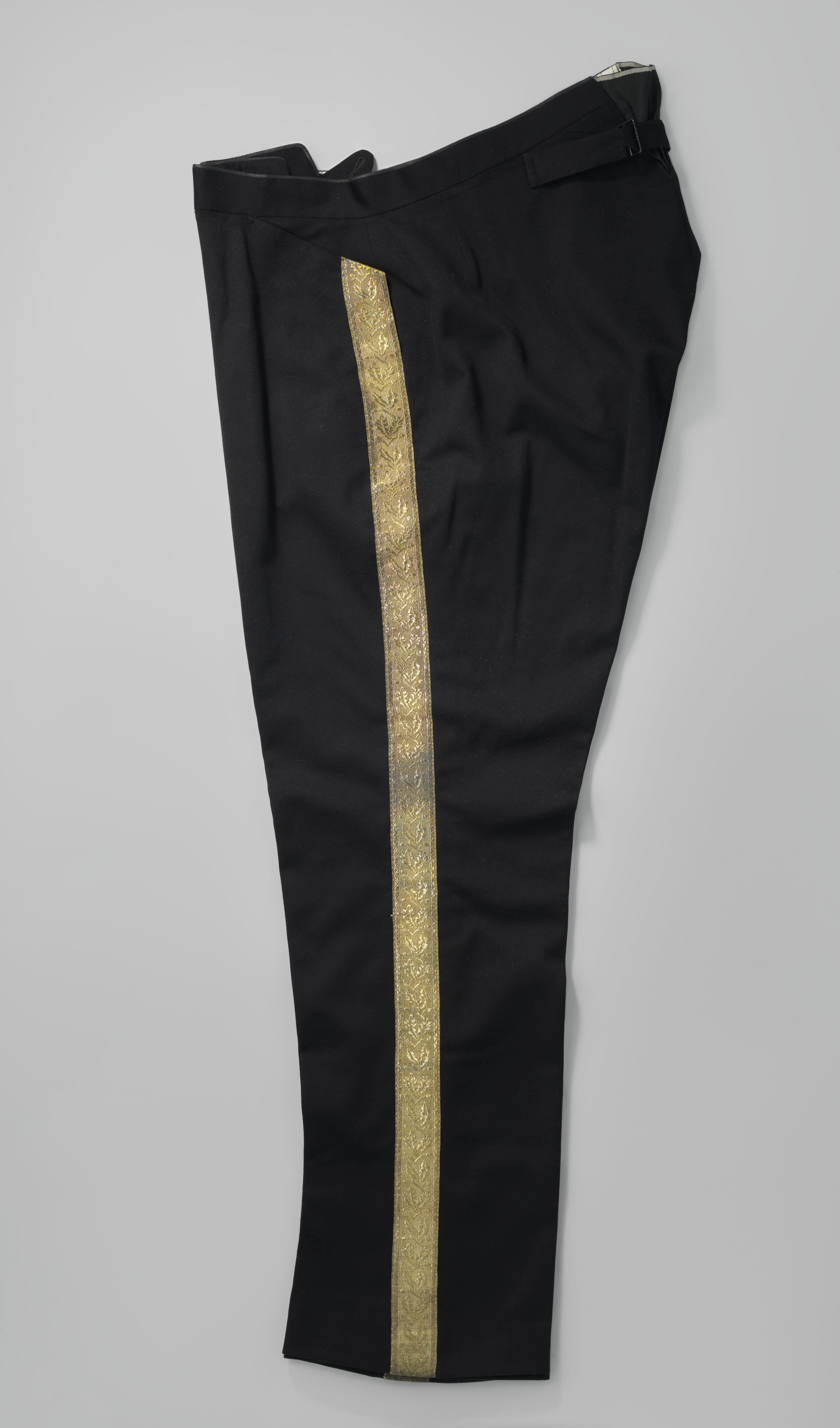 Broek van zwart laken met lange pijpen en opgenaaide tailleband, vanachter met losse banden met een gesp van zwart laken, kleine rechte steekzakken middenvoor