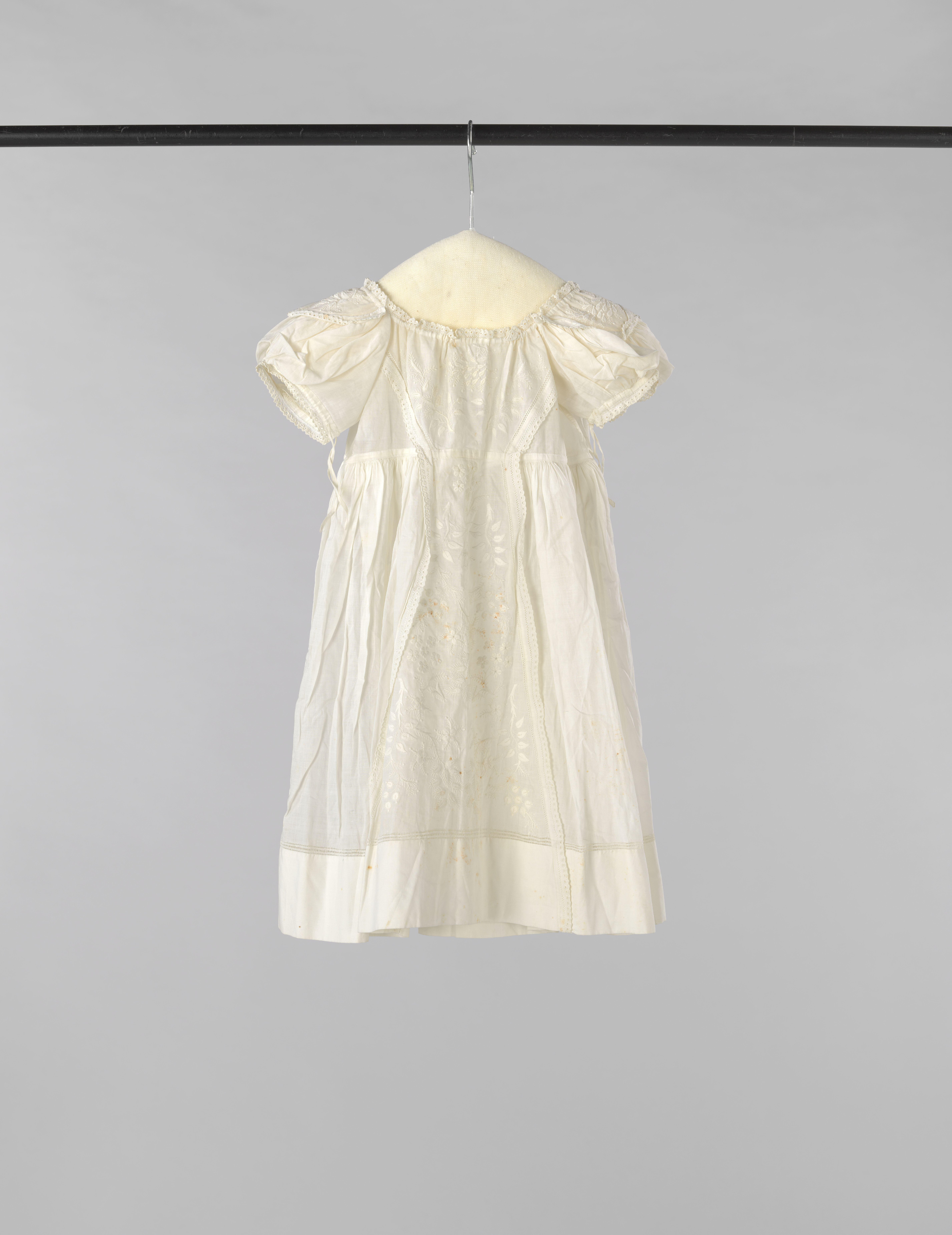 Kinderjurk met hoge taille en achtersluiting met striklinten van witte batist, voor versierd met een breed tussenzetsel plumetis en op de korte pofmouw ook met plumetis.