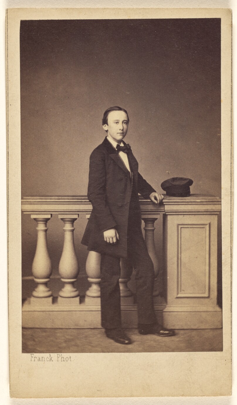 [Unidentified young man standing, his hat resting on a fake railing]