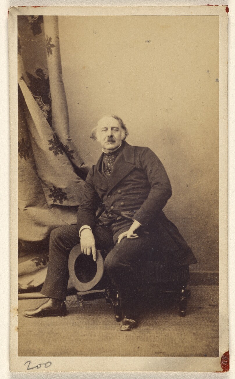 [Unidentified man with longish hair and mustache, seated on hassock, holding hat]