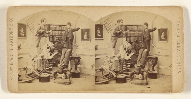 [Group of boys in a living room, one standing on a rocking horse, one standing on a table, one standing on his head, one wearing a hat]