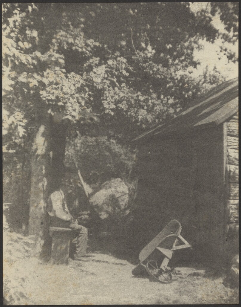 [Old Man Sitting Under Tree by Shed, Wheelbarrow Nearby]