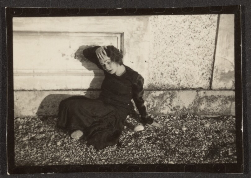 [Contact print from 37mm negative: woman posing on ground against cement wall]