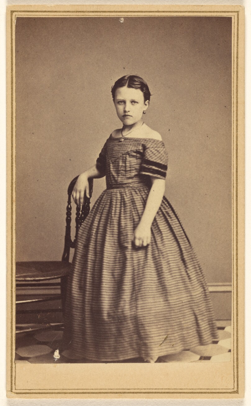 [Unidentified young woman standing, one arm resting on chair back]