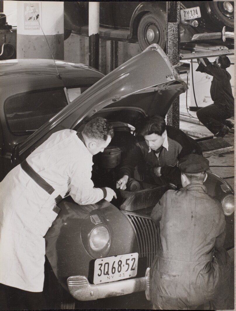 [Three men work under the hood of a car]