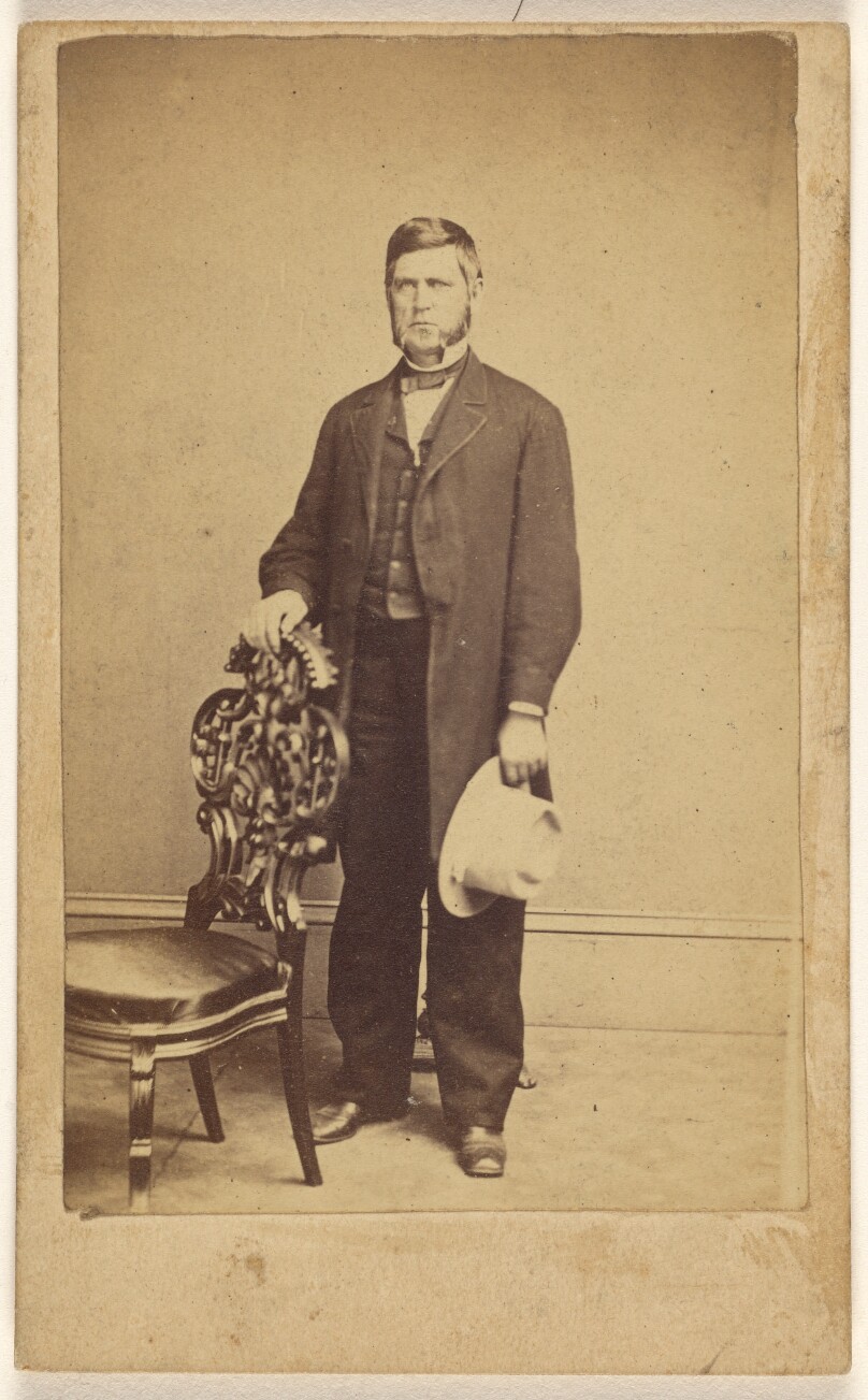 [Unidentified man with long muttonchops, standing, holding a white hat]