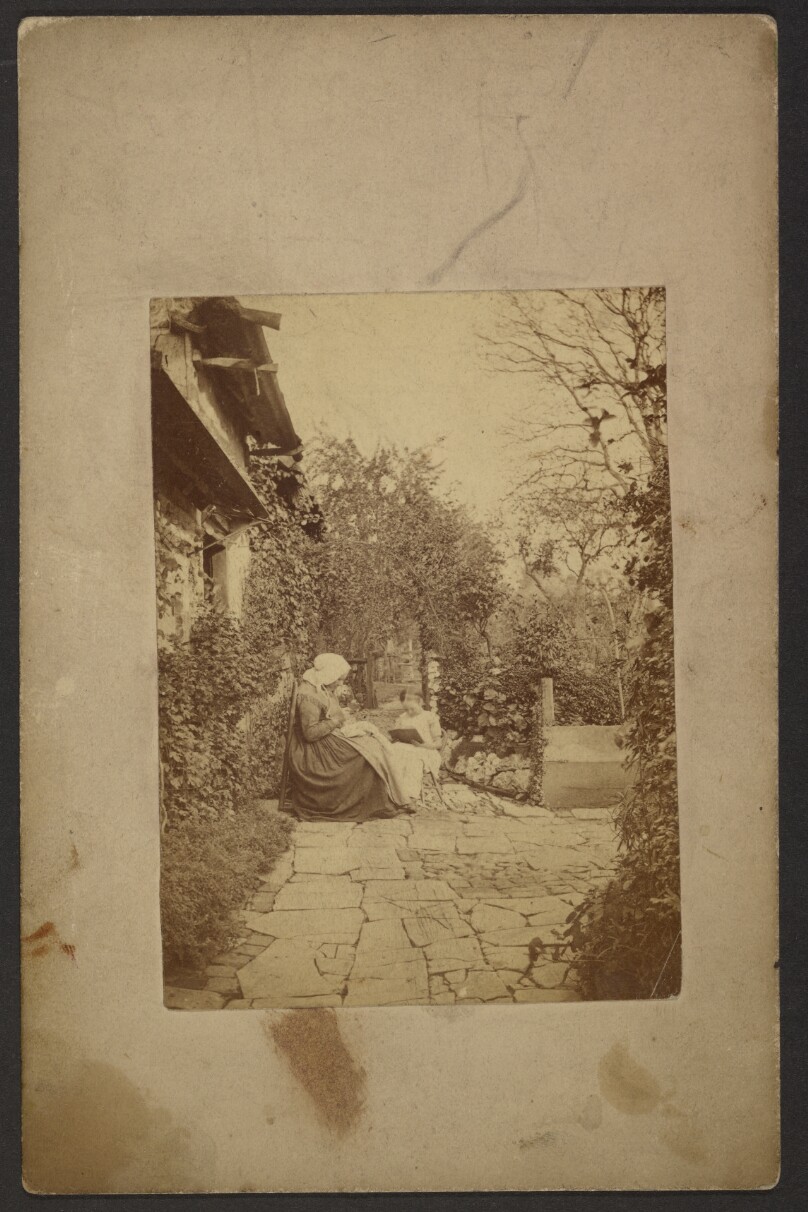 [Woman sewing and young girl reading a book sitting on chairs outside the house]