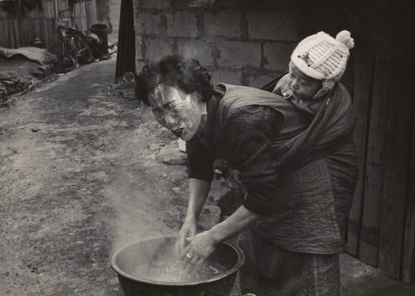Woman Washing Her Face with Suds and Steaming Water While Carrying a Small Child on Her Back