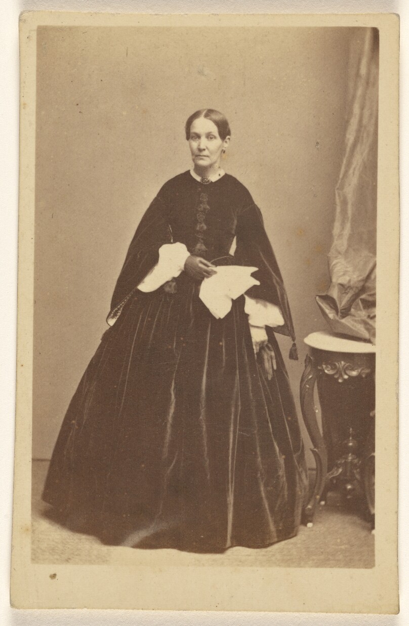 [Unidentified woman wearing a long dark dress, holding a kerchief, standing]