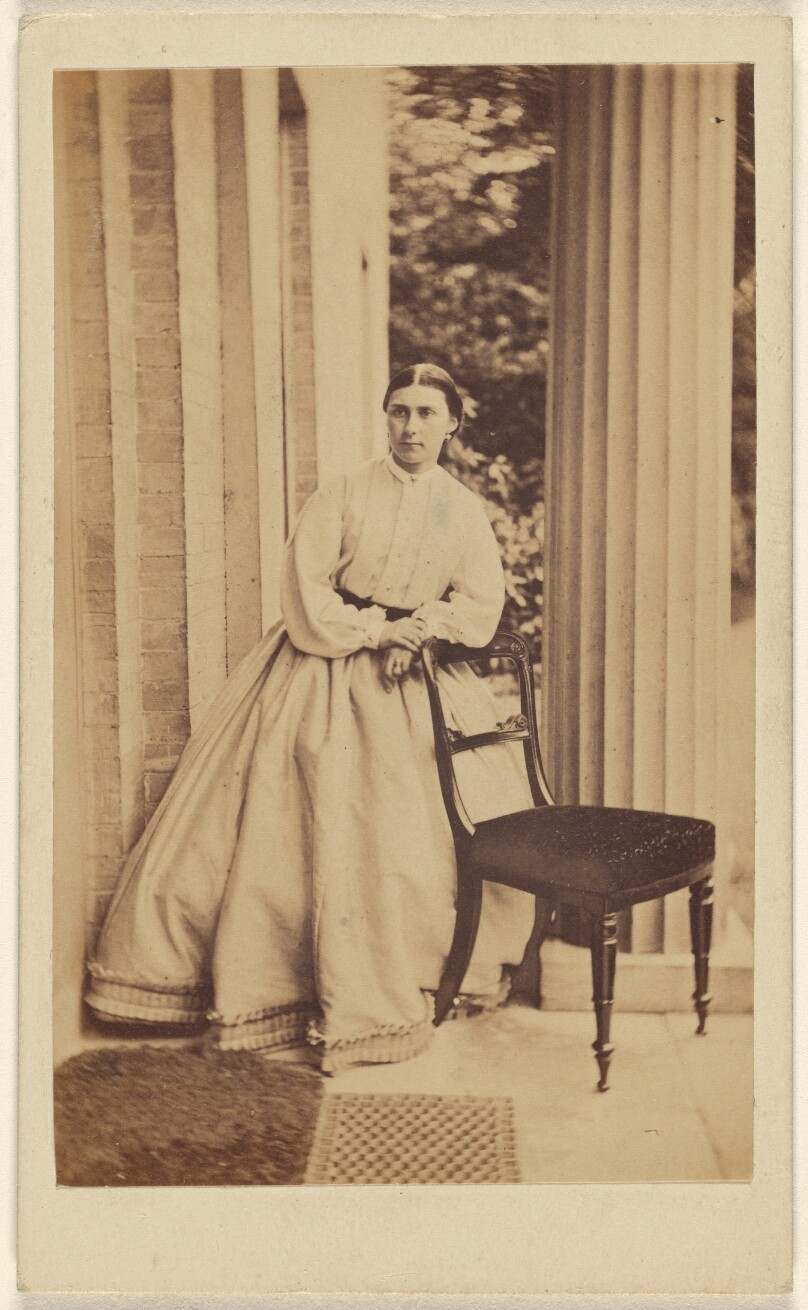 [Unidentified woman standing, leaning on chair back]