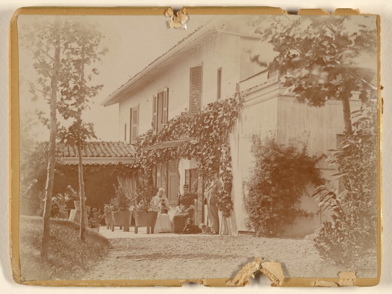 [Group of people with two dogs at front of house, some seated]