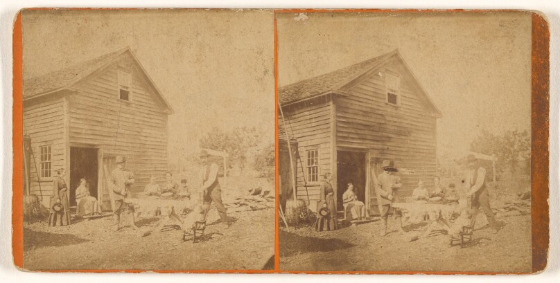 [Group of men and women outside a barn, three women seated at a table]