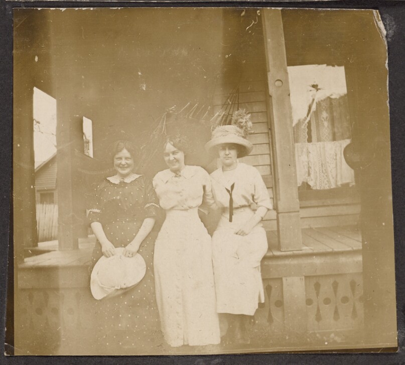[Three women pose on the porch of a house]