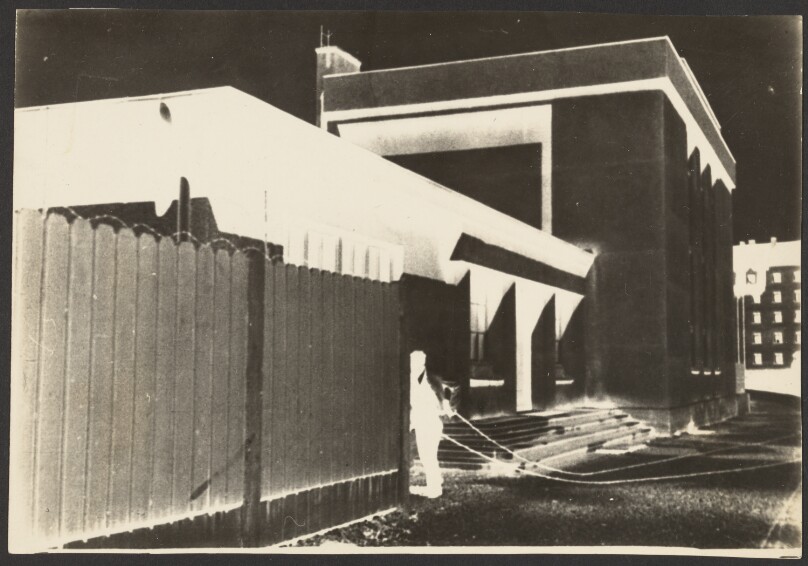[Negative print of a man pulling chain between wooded fence and building]