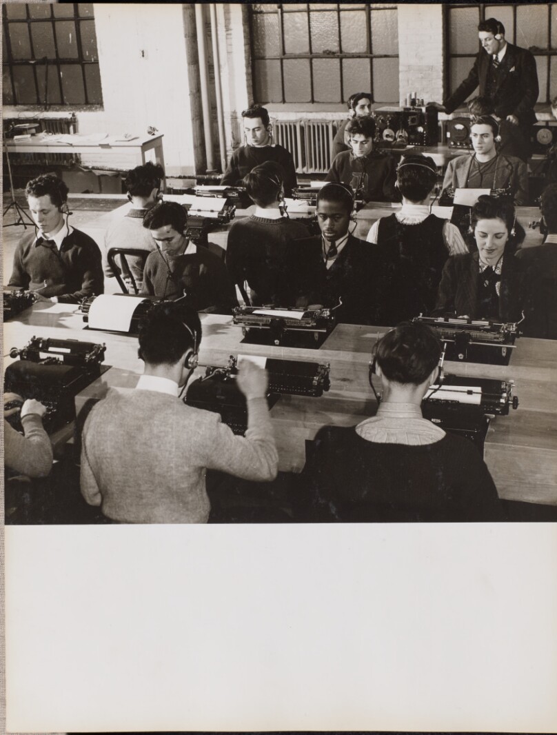 [People sit in front of typewriters while wearing headphones]