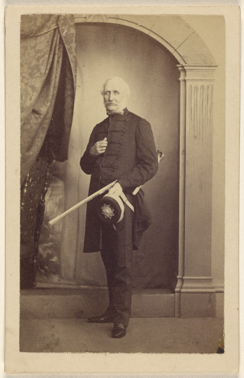 [Unidentified man wearing a dress uniform, standing, holding a hat with his sword under his arm]