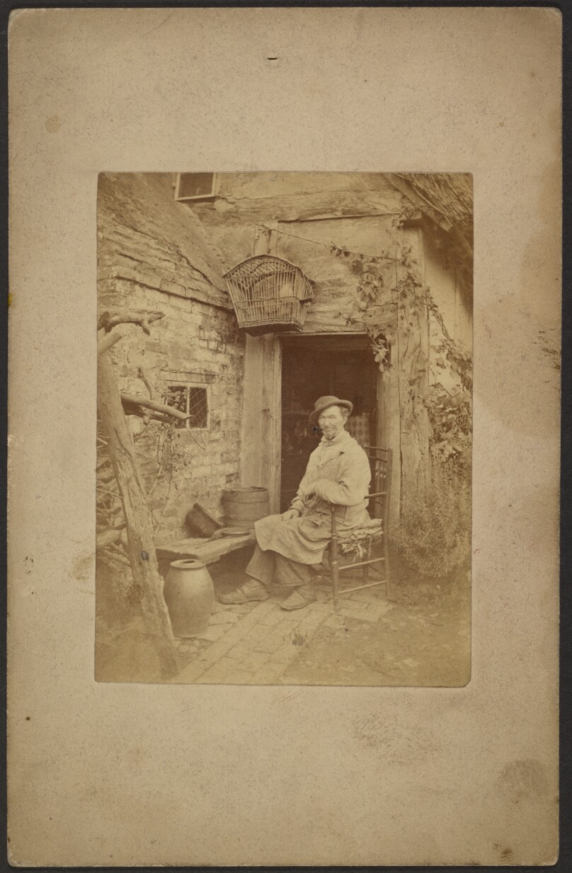 [Man seated outside open doorway, birdcage hanging above]