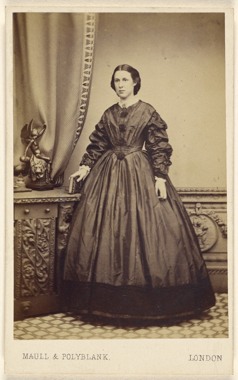 [Unidentified woman wearing a long dress, holding a book atop a credenza, standing]