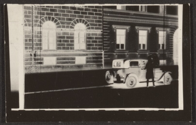 [Contact print from 37mm positive film: woman before car in front of a brick building facade in negative]