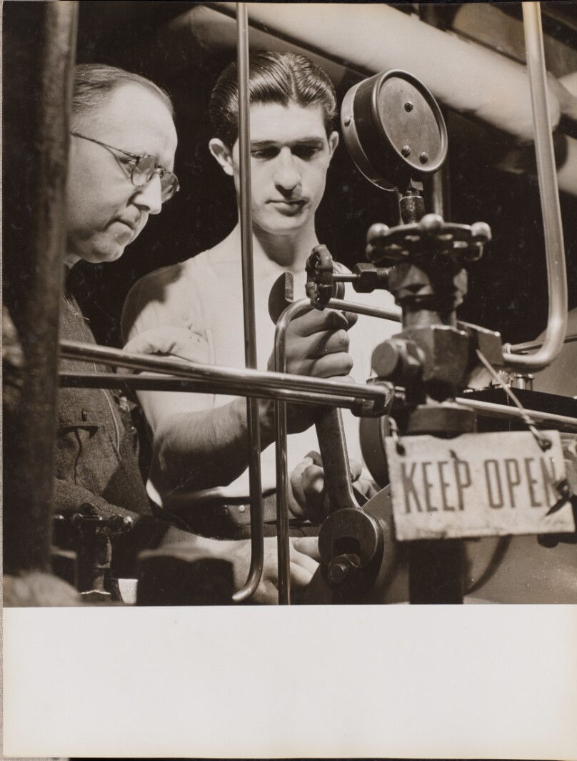 [Two men adjust a tank with a large wrench]