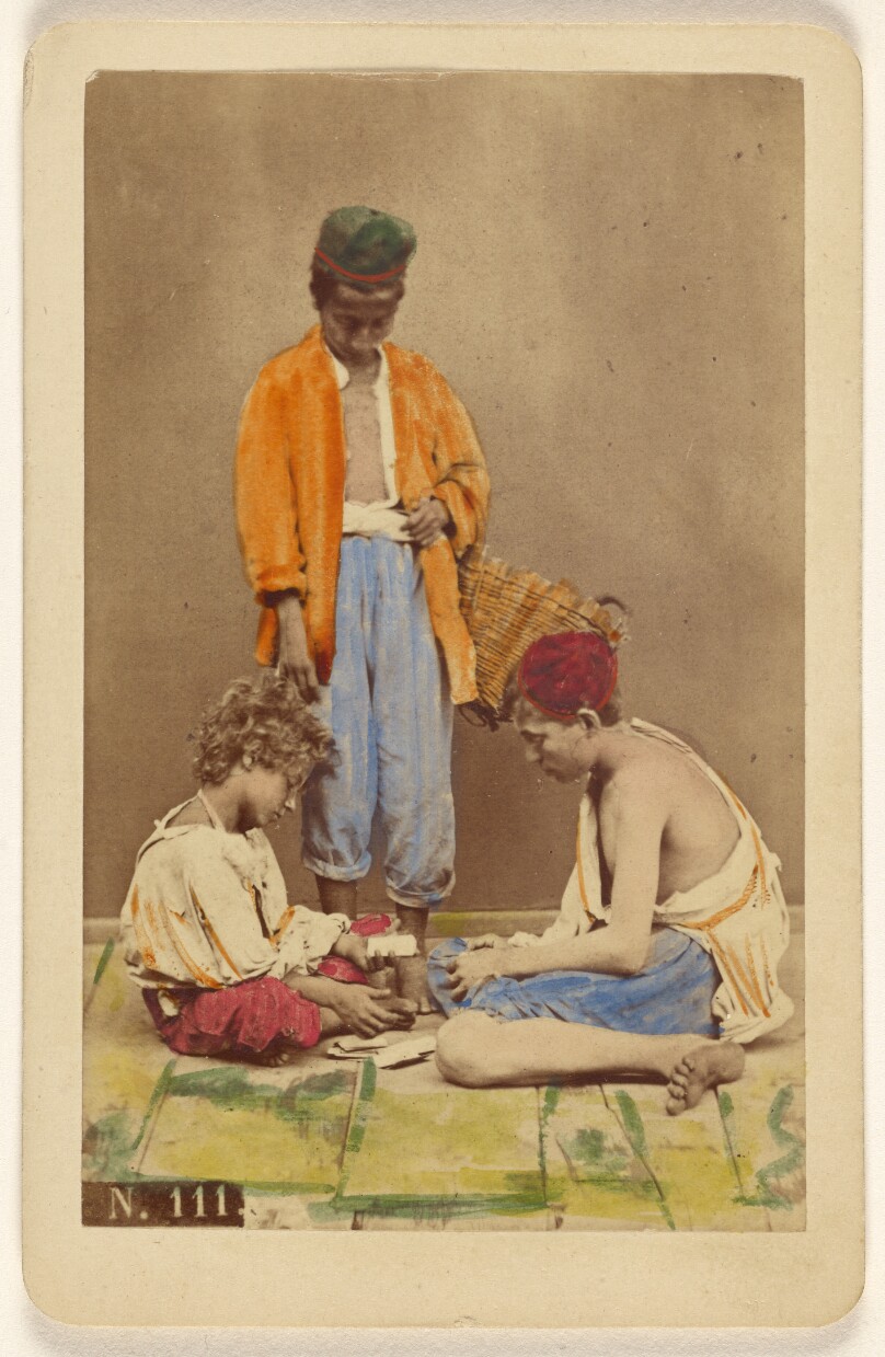 [Unidentified man with basket standing, plus two boys seated playing cards]
