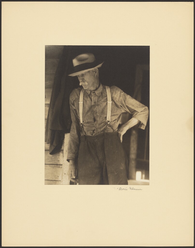 [Mustached Old Man with Straw Hat and Suspenders Standing in Doorway]