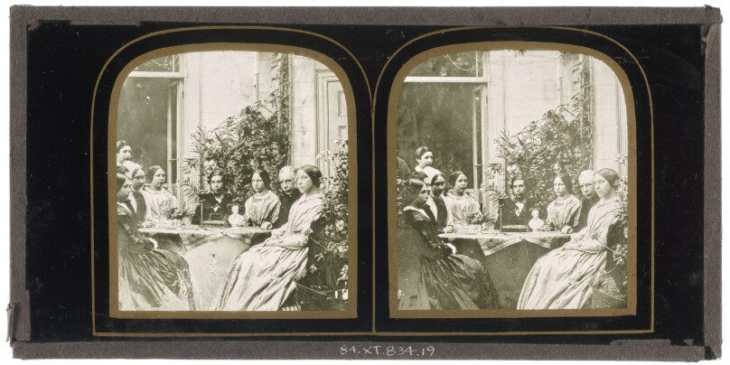 [Group of unidentified British men and women seated around a table with a small bust of a man at center]