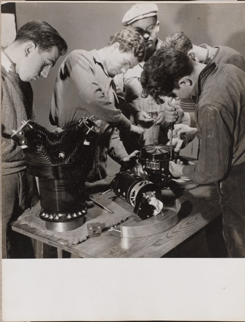 [Five young men work around a work bench]