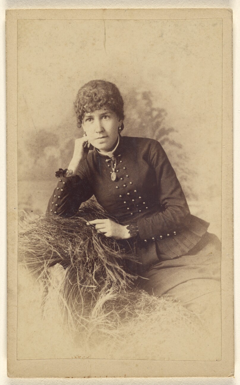 [Unidentified woman seated with hand on cheek, posed on a pile of hay]
