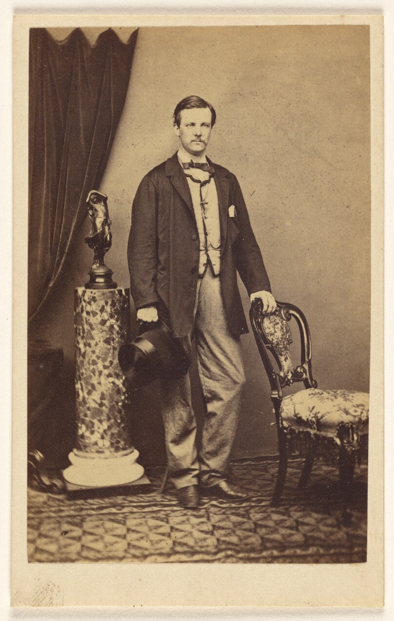 [Unidentified man with moustache standing, holding top hat in one hand, the other hand on chair back]