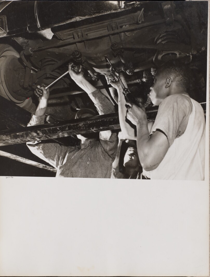 [Three men work on the underside of an automobile]