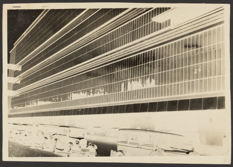 [Negative print of glass-windowed facade of building with parked cars]