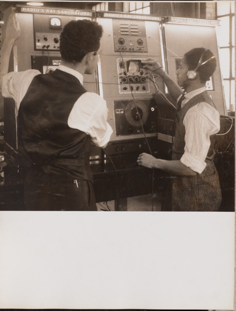 [Two men wearing vests work with large machines]