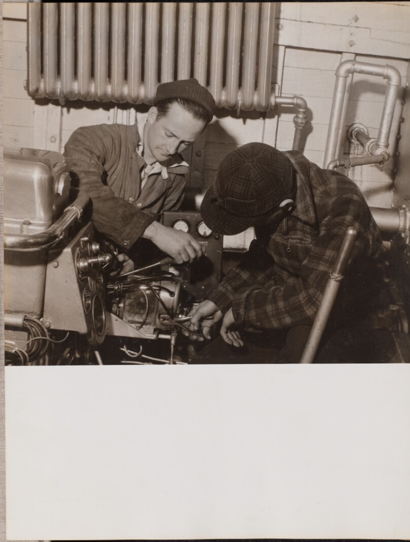 [Two men work on a machine using a screwdriver and a wrench]