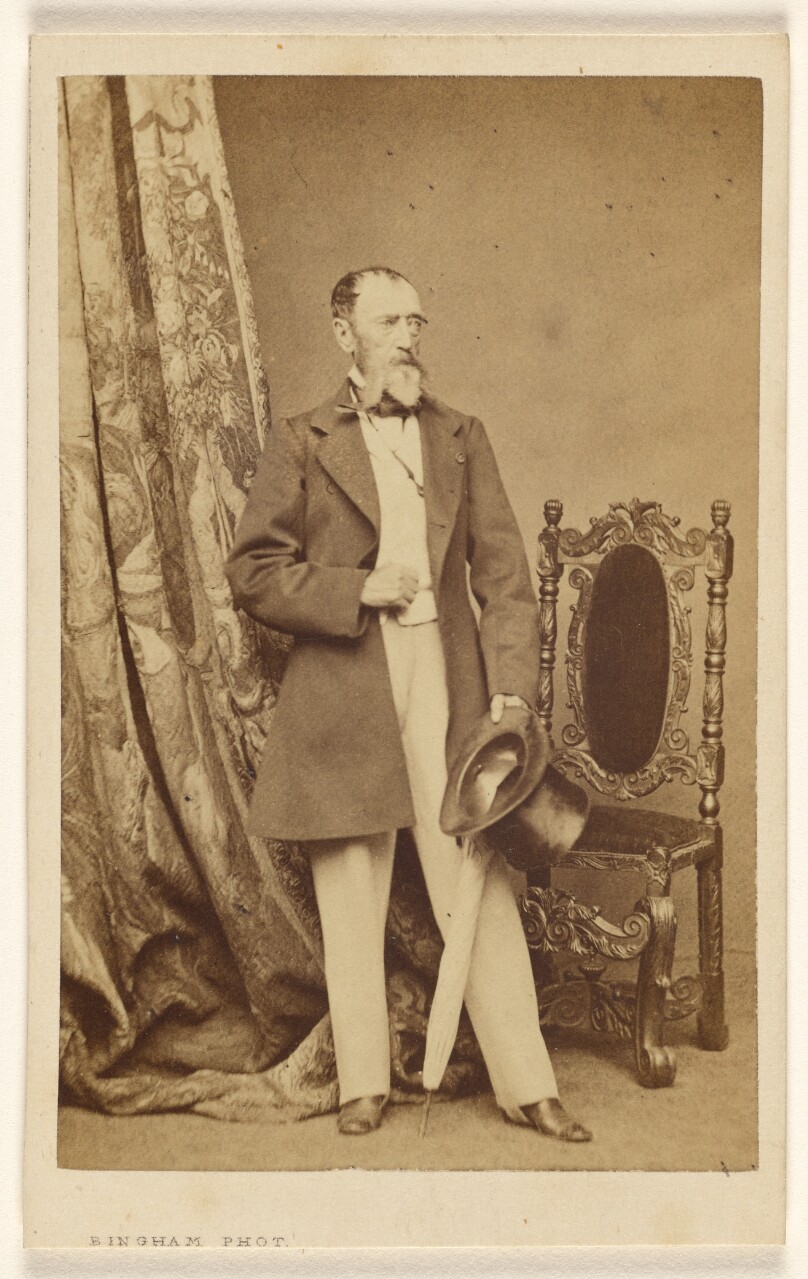 [Unidentified man with full white beard, standing, holding a top hat and umbrella in his left hand]