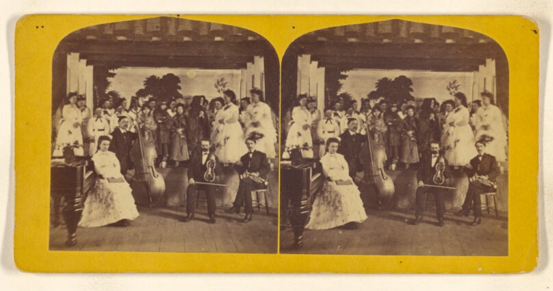 [Cast of a play on stage, three male musicians seated in front with well-dressed woman]