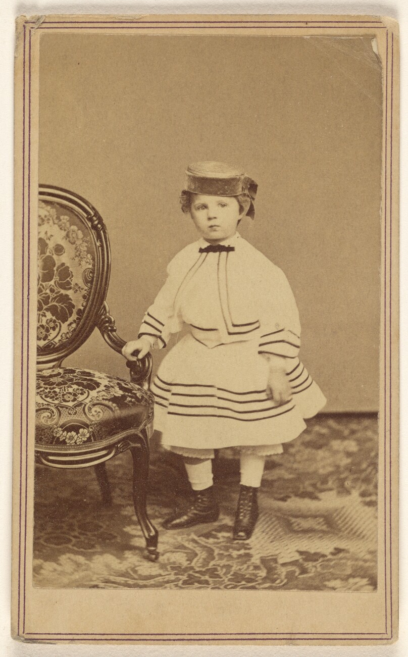 [Unidentified little girl wearing a pill-box hat, standing, holding the arm of a chair]