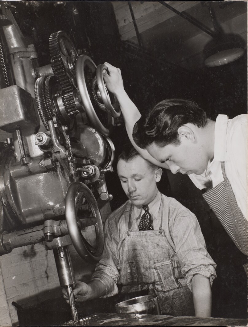 [Two men look at machinery with a drill bit]