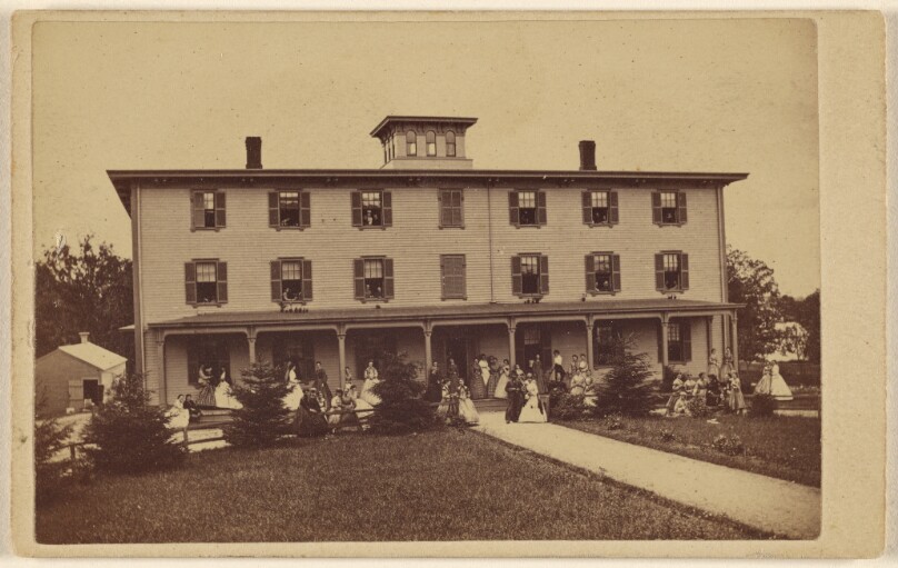 [View of a house with numerous people on porch and lawn]