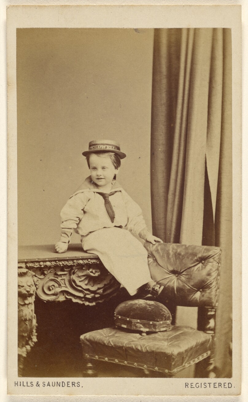 [Unidentified little boy seated on a table, wearing a straw hat with a "Speedwell" banner]