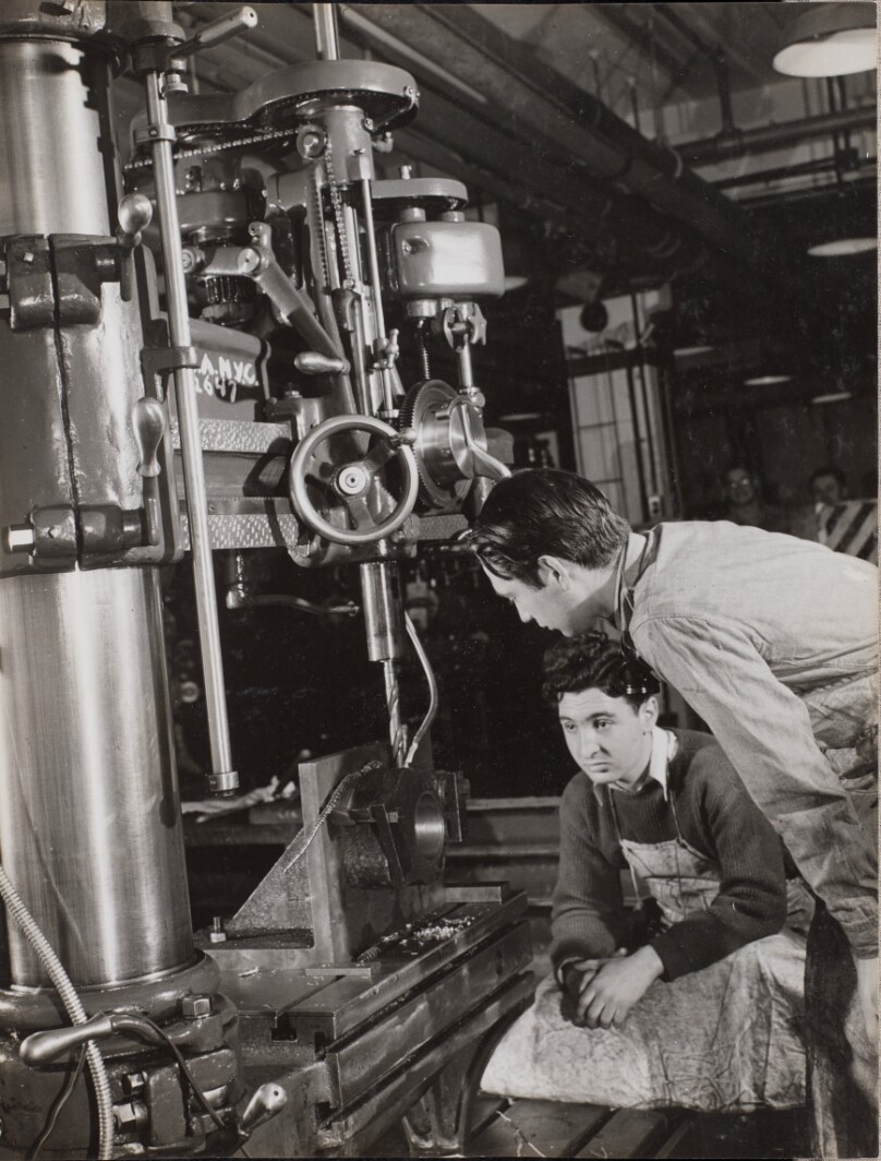 [Two men sit near a large piece of machinery with a drill bit attached]